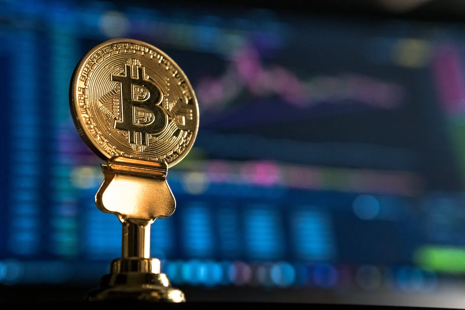 A gold-colored bitcoin coin on a small pedestal in front of a blurred price chart